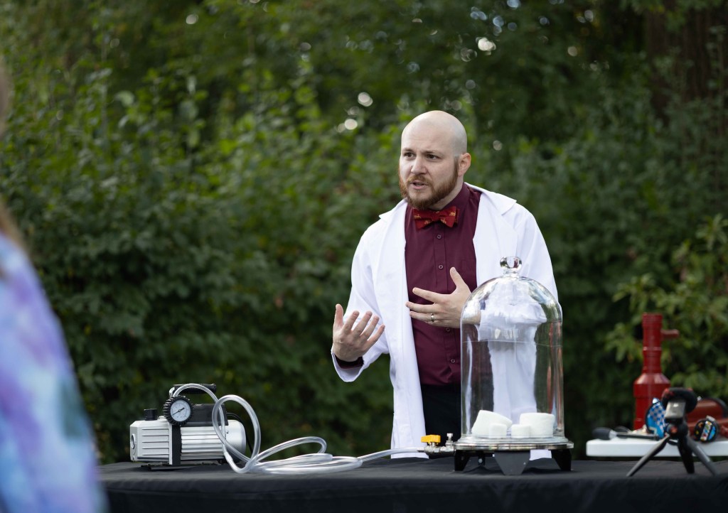A scientist in a lab coat and bow tie gestures while explaining a demonstration, with scientific equipment visible on a table in a green outdoor setting.
