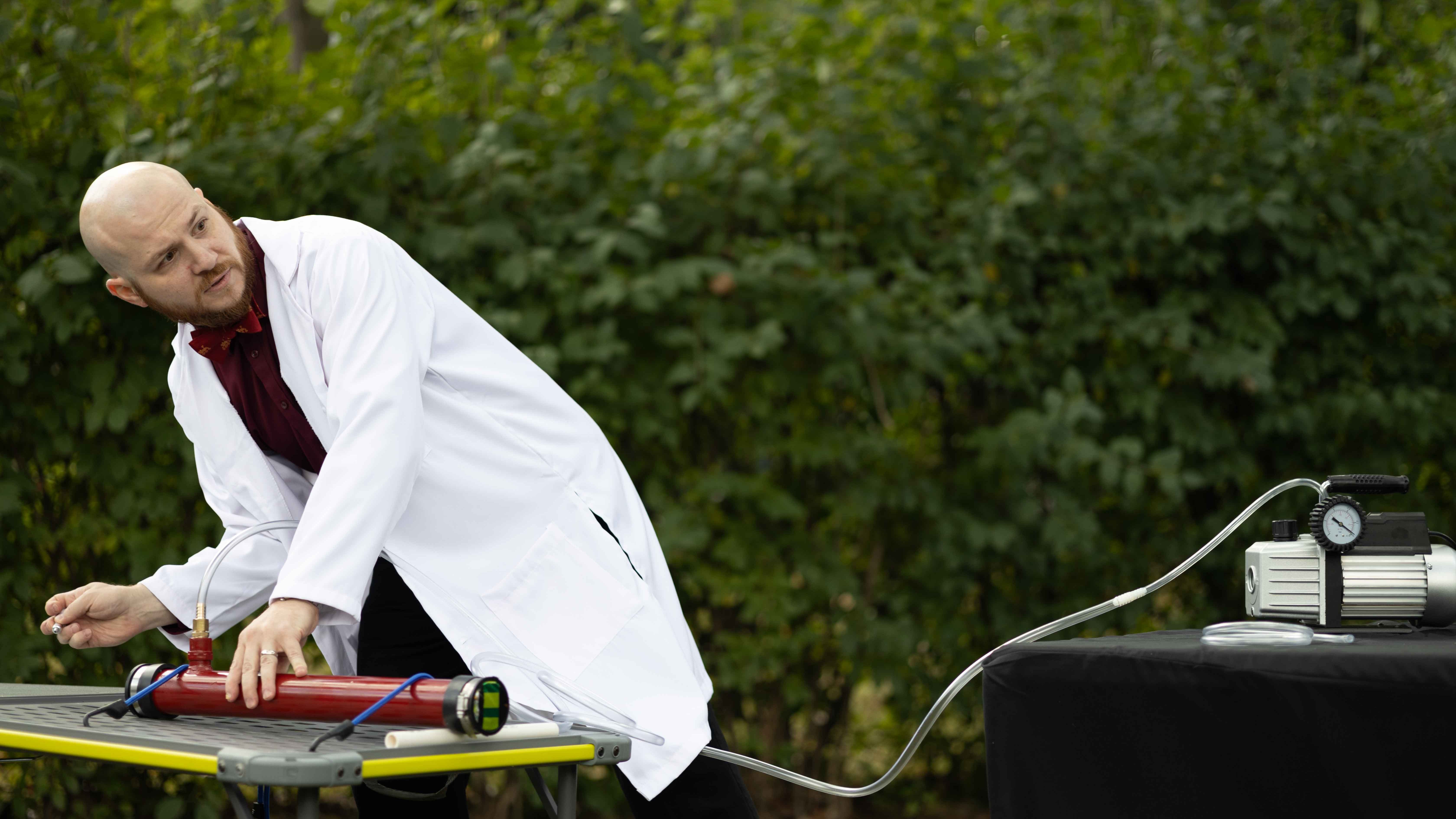 A person in a lab coat is operating scientific equipment on a table outdoors, with a pressure gauge and pump visible in the background.