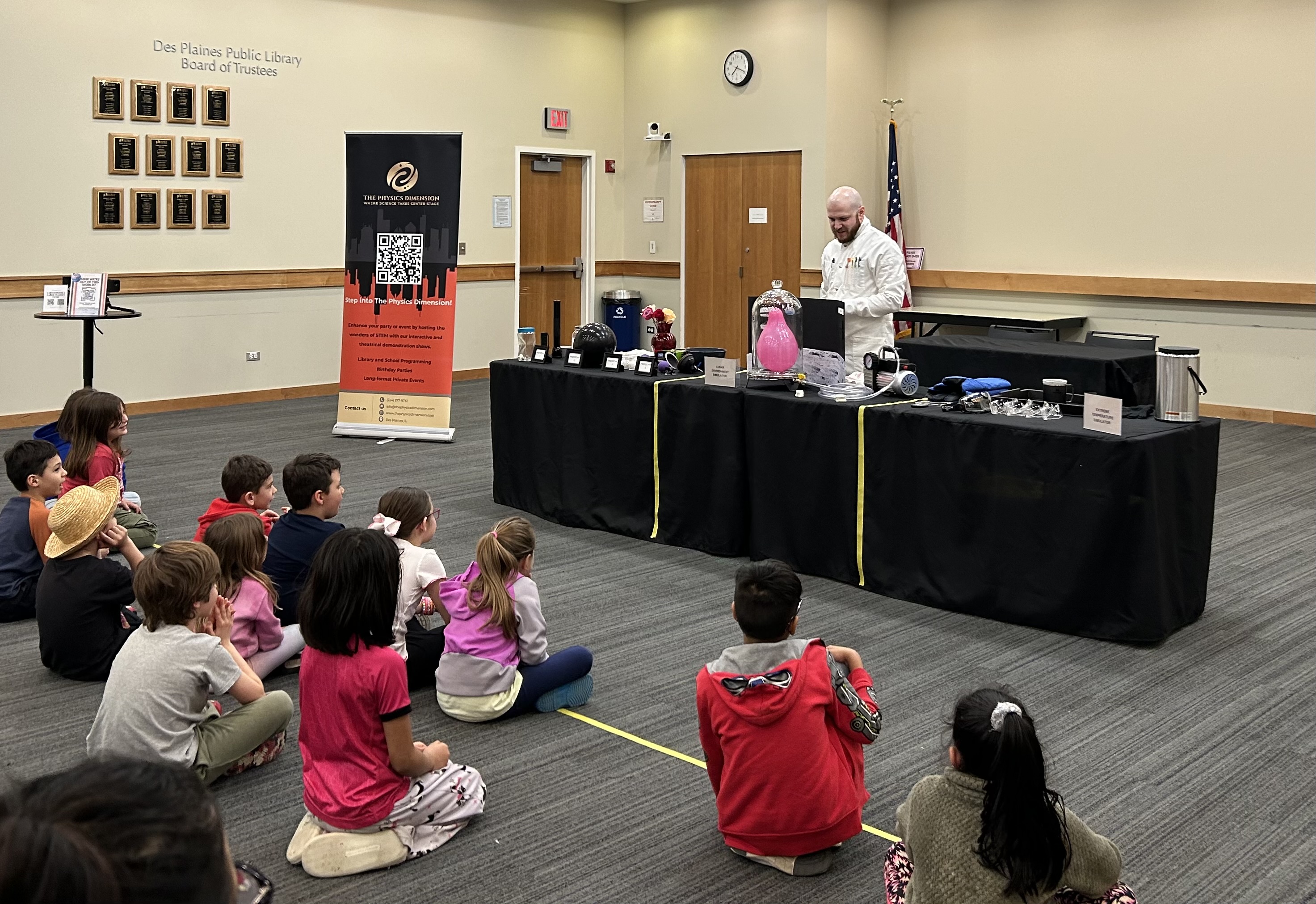 A science demonstration at Des Plaines Public Library featuring a presenter in a lab coat engaging with a group of children. The presenter is using various equipment and materials displayed on a table covered with a black cloth.