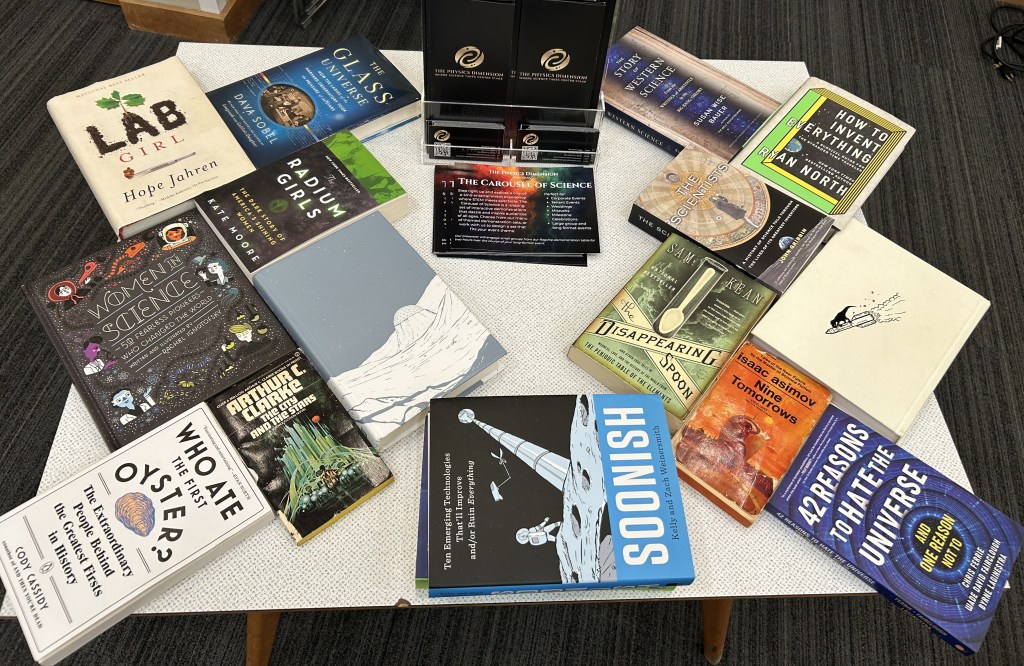 A collection of science-related books displayed on a table, featuring titles like 'Lab Girl', 'Women in Science', 'The Radium Girls', and 'How to Invent Everything'.