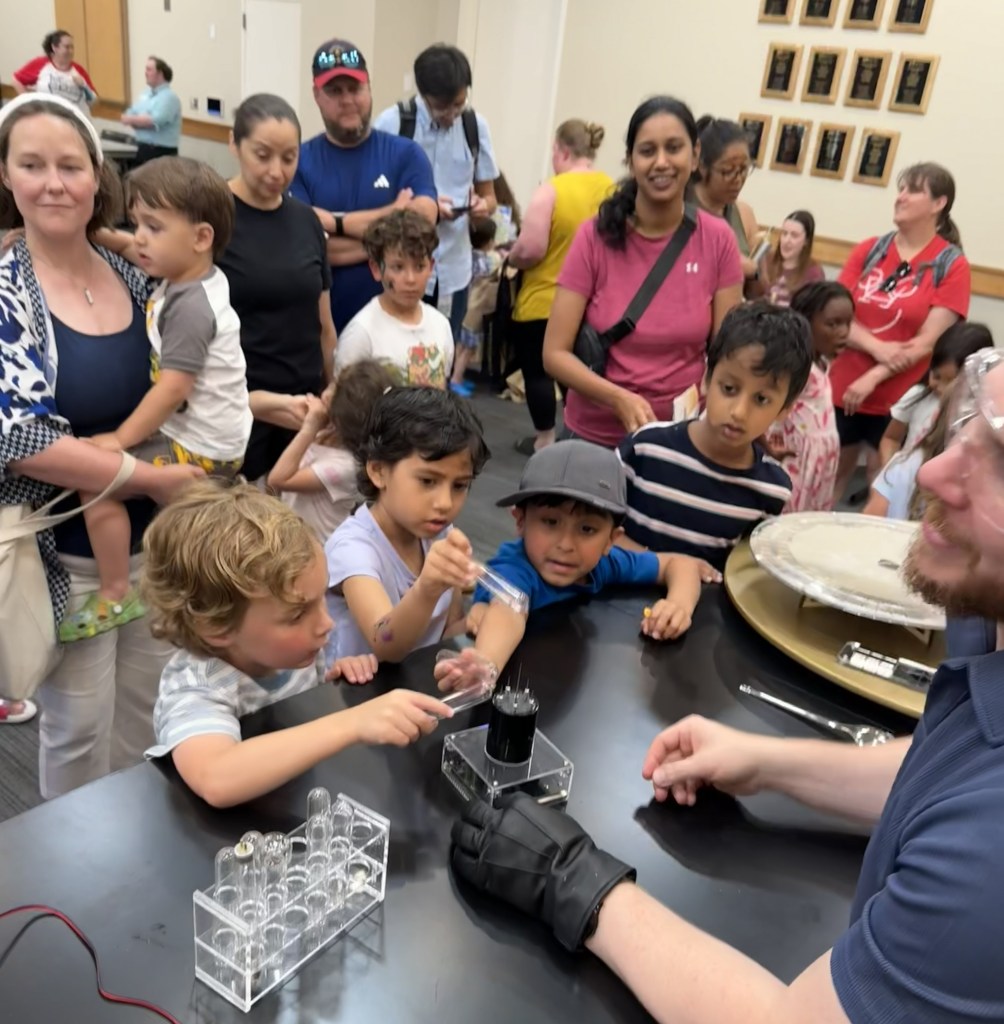 A group of children and adults gathered around a table, watching a demonstration with scientific equipment. One child is pointing at a piece of apparatus as an adult in gloves explains it.