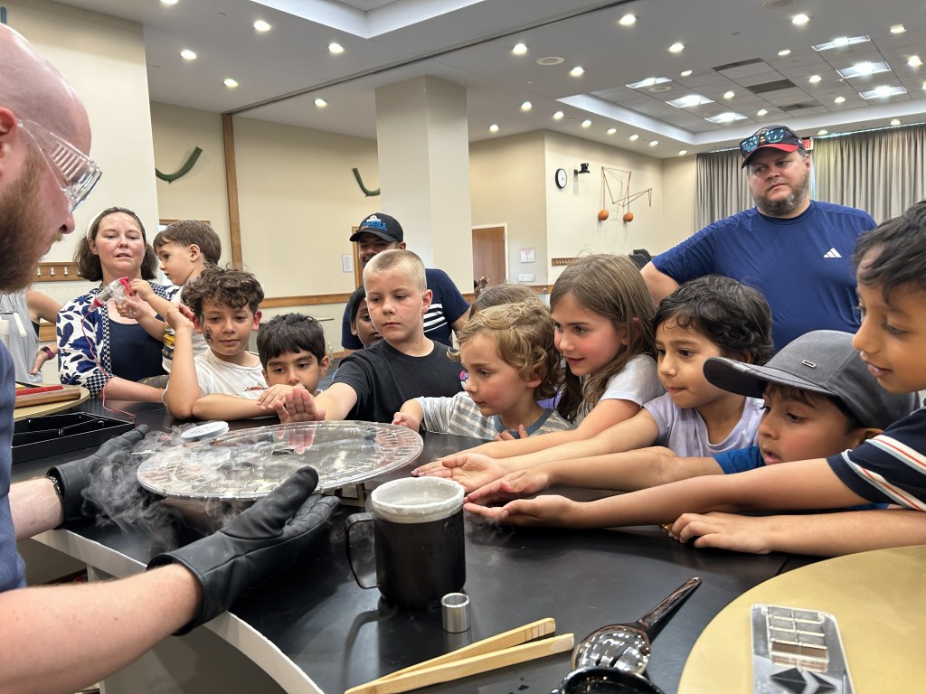 A group of children and adults are gathered around a table, watching a demonstration involving dry ice and a circular dish, with steam or fog visible around the edges. The children are eagerly reaching towards the dish, showcasing their curiosity and excitement.